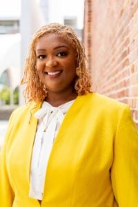 Headshot of Ashley Stepps in a yellow blazer and white shirt on an outdoor brick wall background.