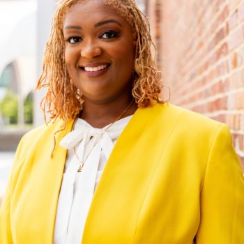 Headshot of Ashley Stepps in a yellow blazer and white shirt on an outdoor brick wall background.